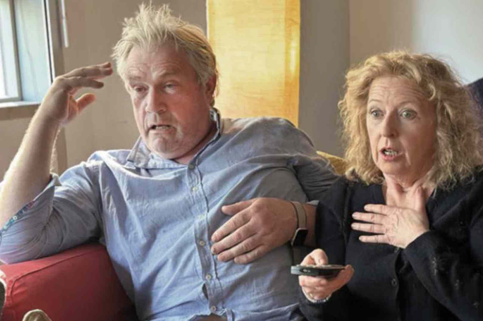 Man and woman sat close together on a sofa, both looking ahead with shocked expression, the woman is holding up a remote control.
