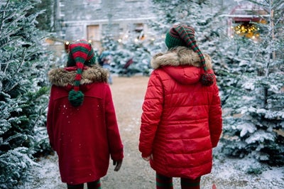 Rear view of 2 children in red coats walking with snow covered christmas trees
