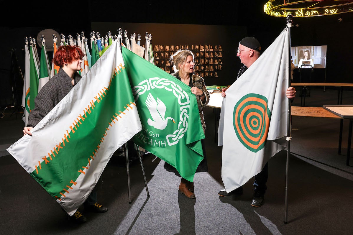 3 people in a discussion, each holding a large flag on a pole with a large number of flags behind