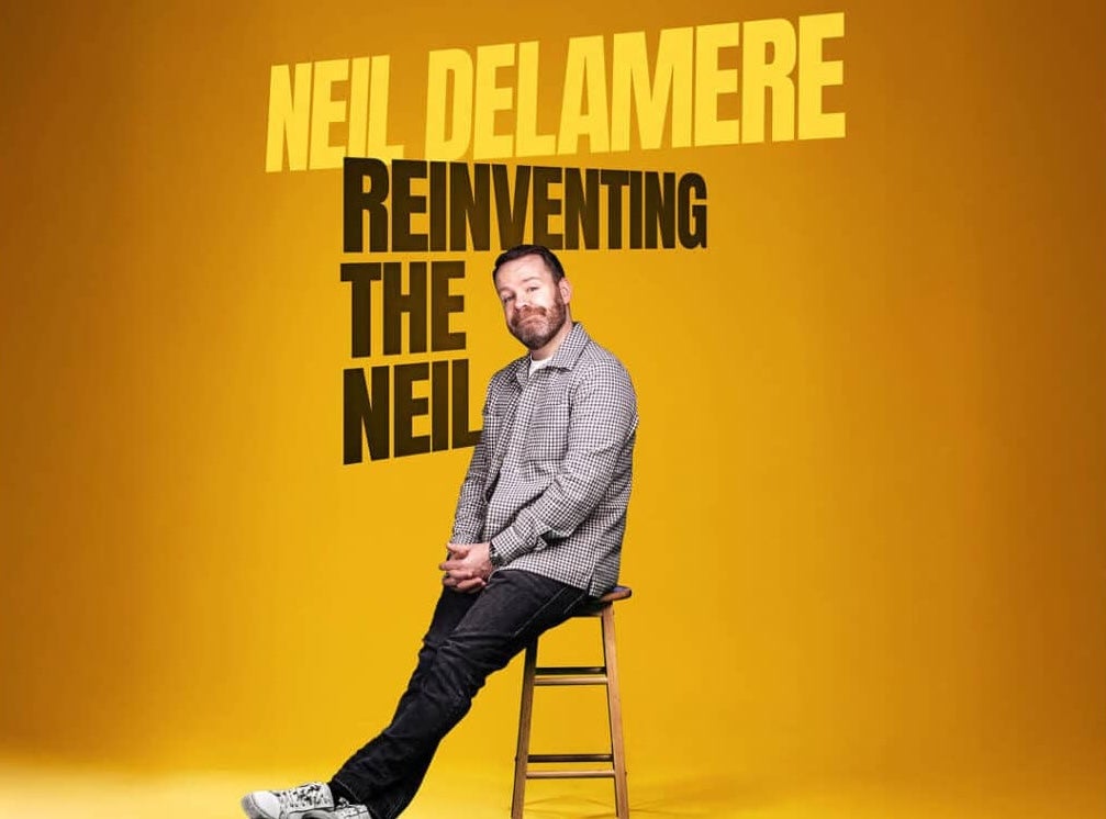 Award-winning Irish comedian Neil Delamere, a smiling man perched on the edge of a tall stool.