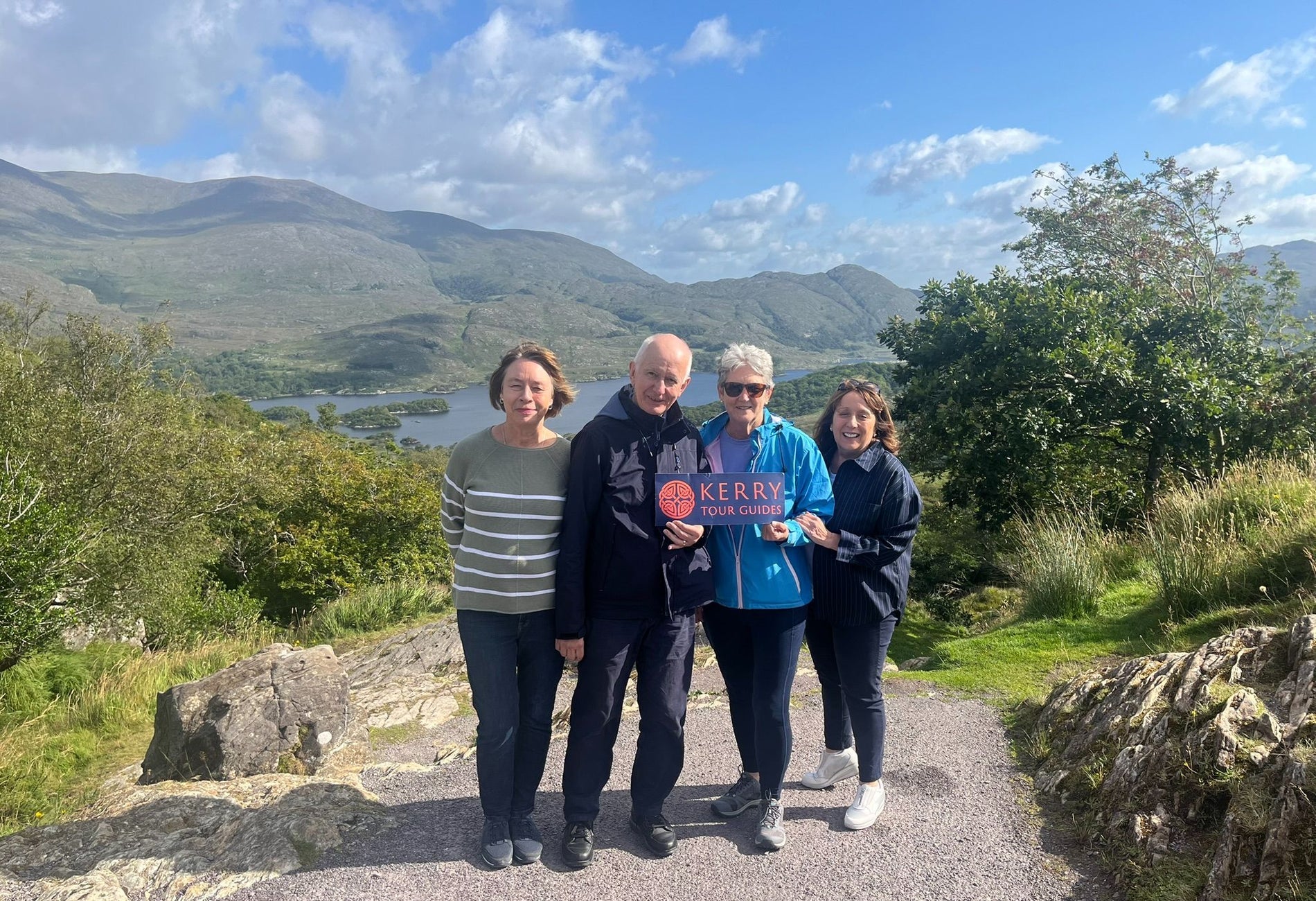 Four people on a path holding a sign with lakes and mountains in the background