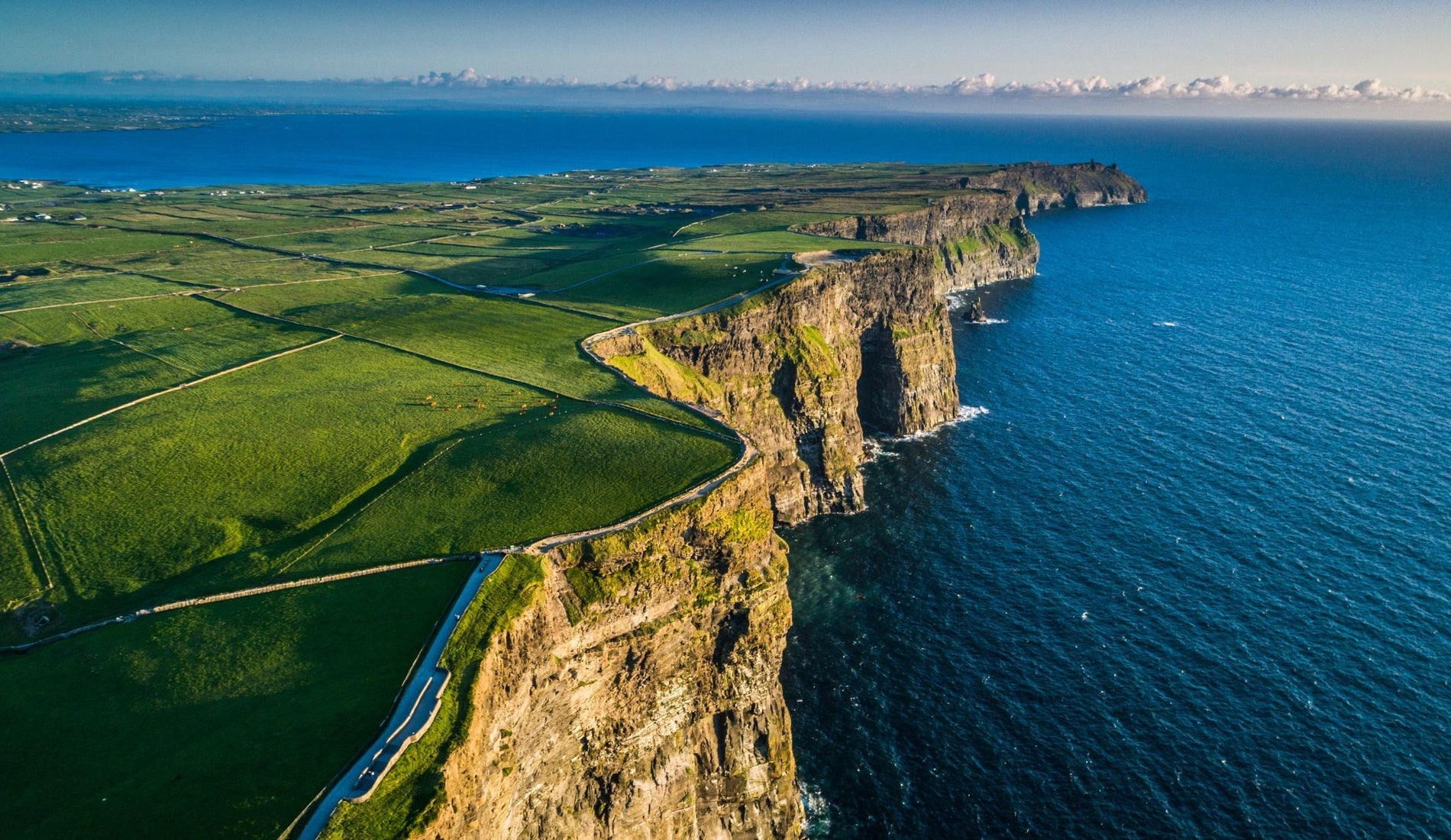 Steep coastal sea cliffs overlook the Atlantic Ocean bordered by green fields and walking paths