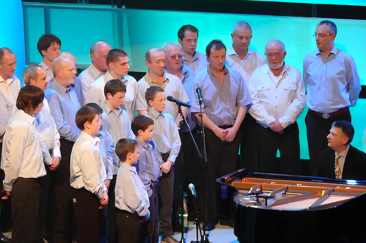 A large group of men and boys all wearing pale, plain coloured shirts, gathered around a man seated at a grand piano.