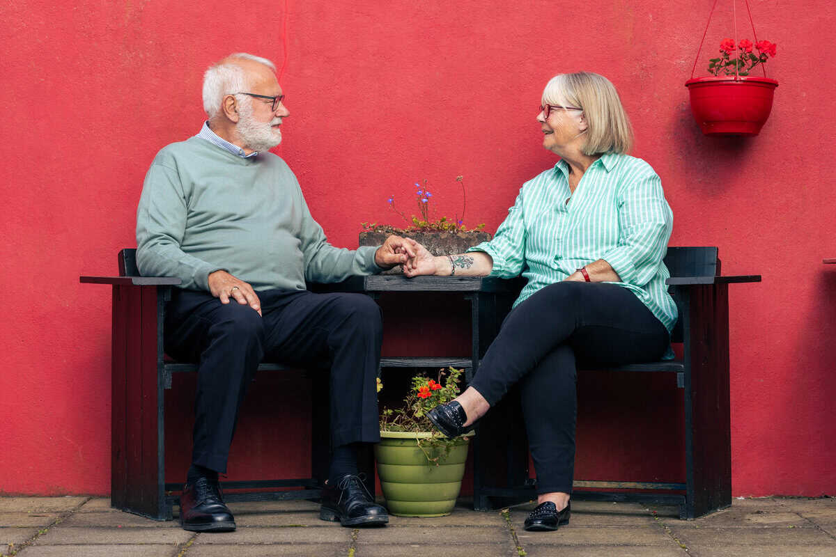 A man and woman seated on dark seats looking at each other, holding hands.