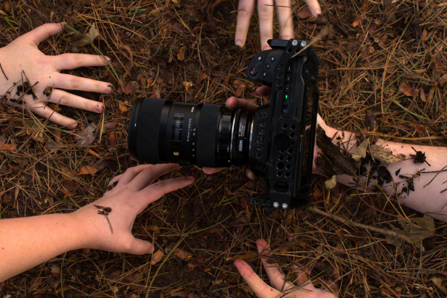 Looking down at 5 hands reaching for a camera with large lens in brown earth and twigs.