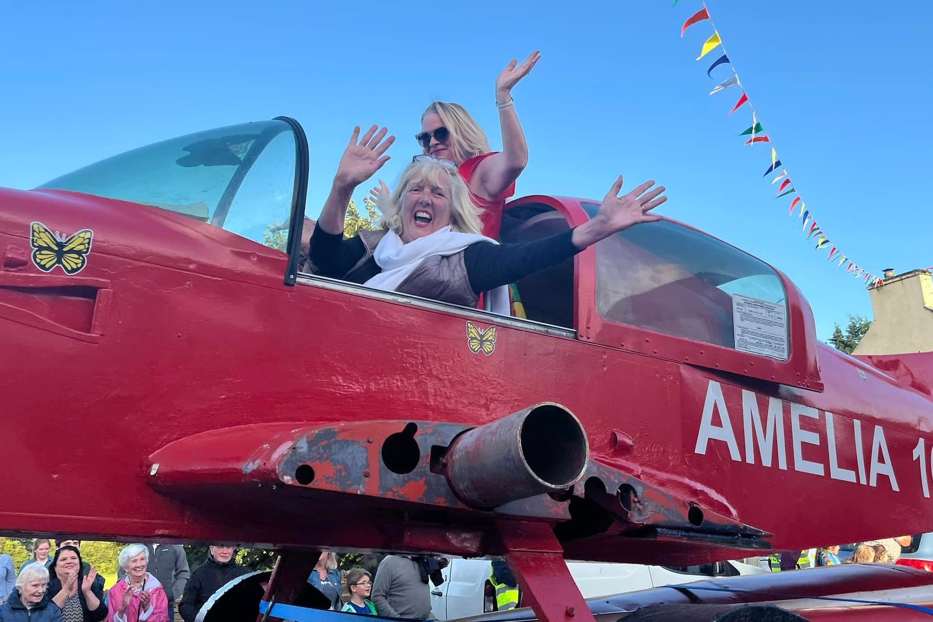 2 smiling and waving women in the cockpit of an old red small aeroplane