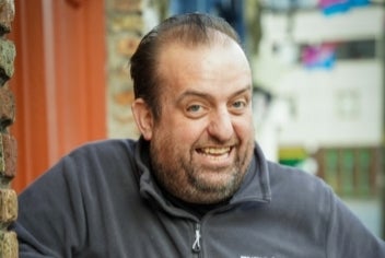 A smiling man in grey fleece is leaning on a window ledge outdoors.