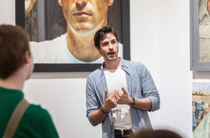 A man is talking and gesturing with his hands, standing in front of pictures on a wall.