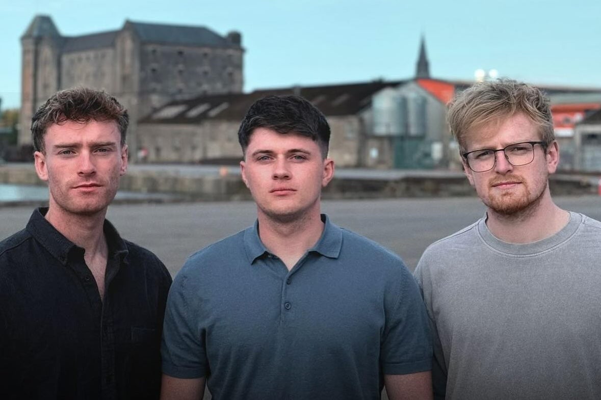 3 men standing close together with water and buildings in the background.
