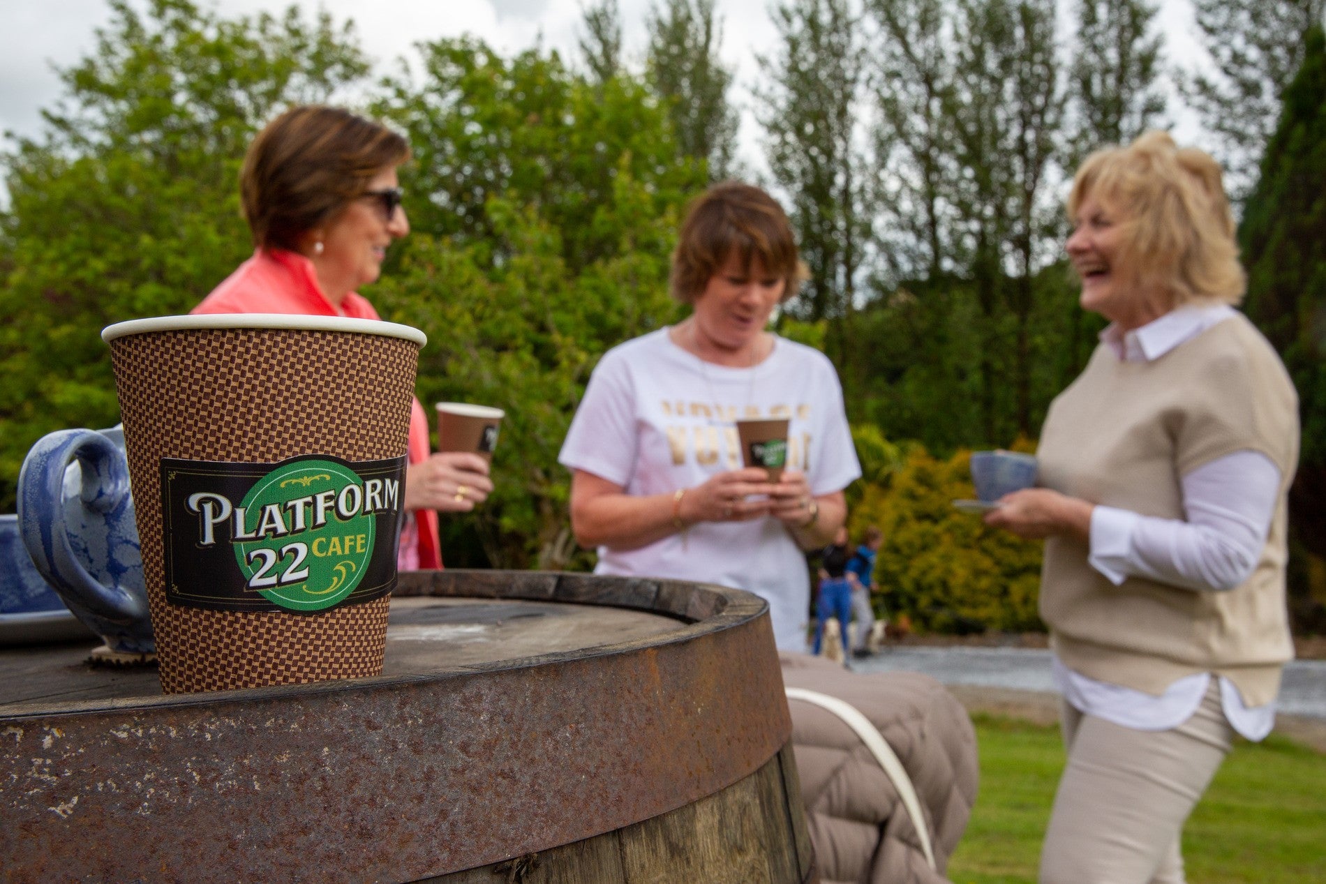 Three ladies outside having coffee with a take away coffee cup on a barrel to the forefront