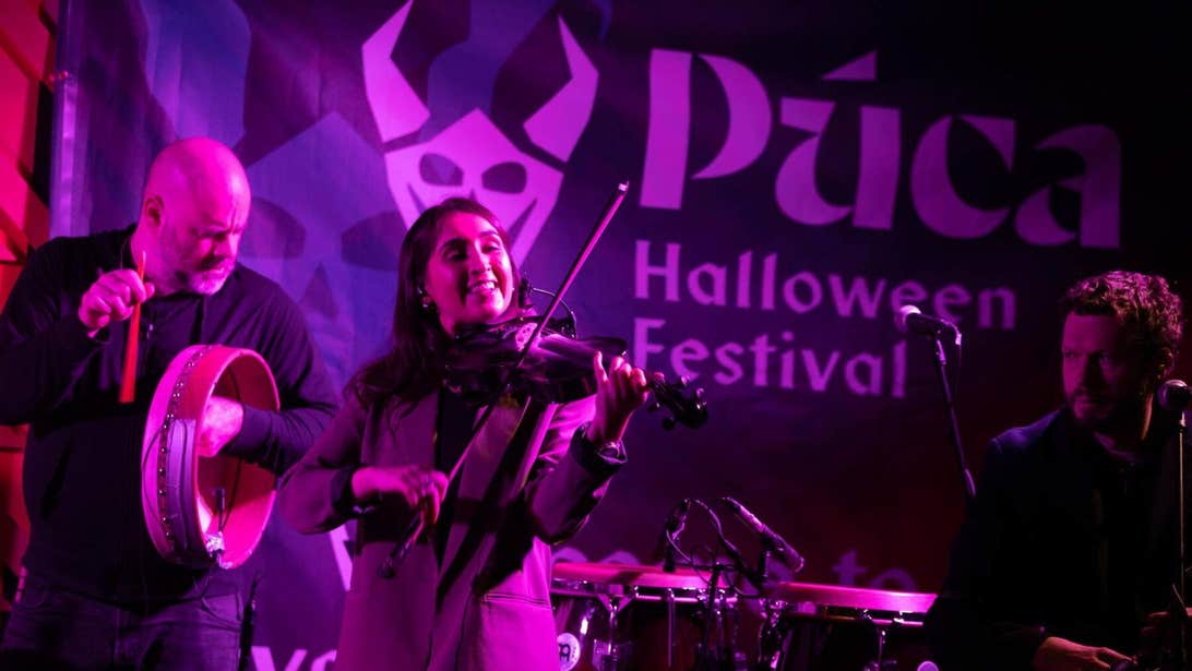 Three musicians on stage at the Púca Festival in Meath. A woman is playing a violin, a man is playing a drum, and the other man is sitting at a microphone.