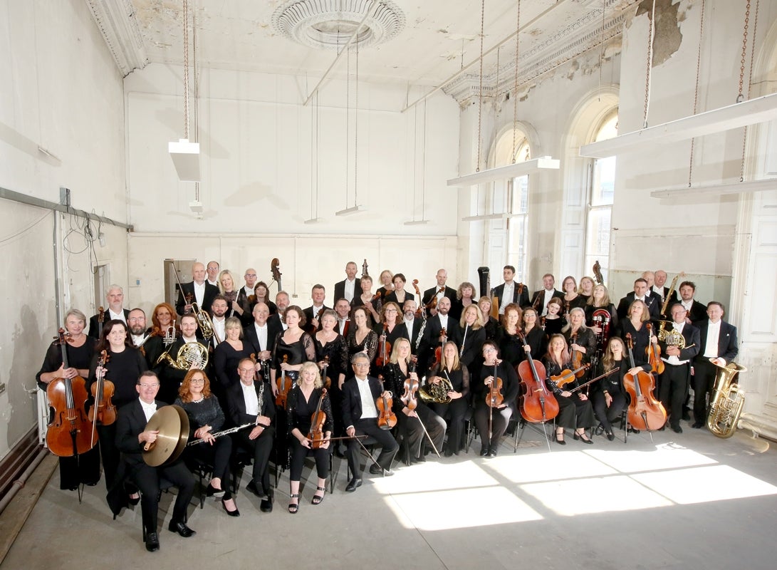 National Symphony Orchestra Ireland, a large orchestra posing in a group with their instruments in a large white room.