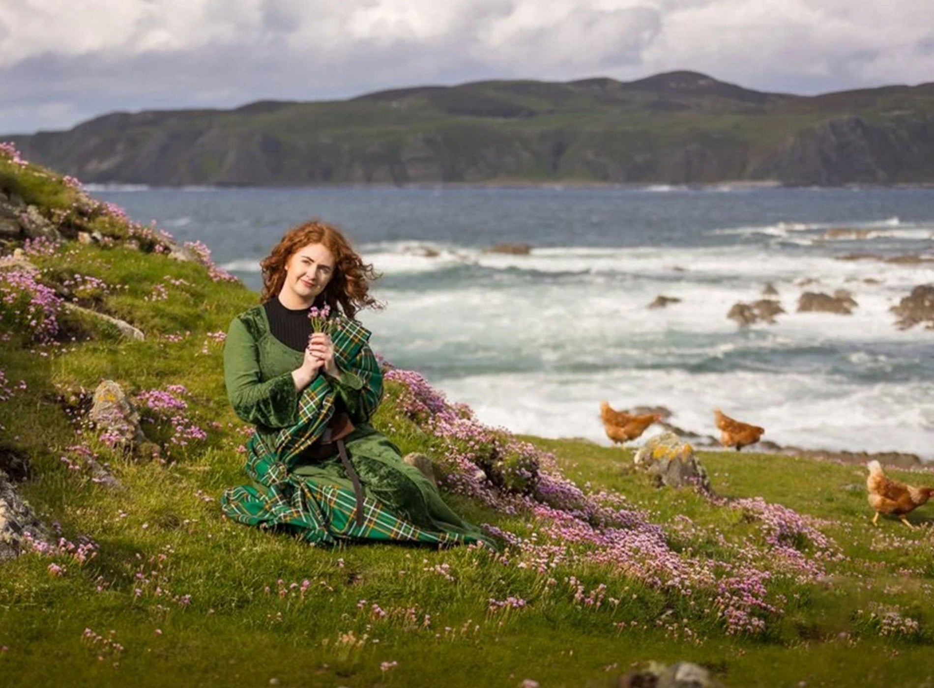 Person dressed in green tartan sitting among pink wildflowers on a grassy hillside near the sea