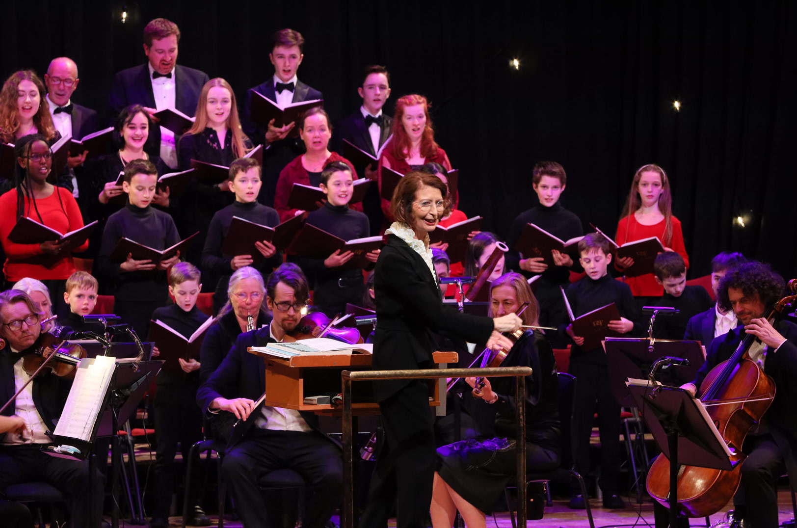 A choir of adults and children with some people playing stringed instruments sat at the front, a conductor is turned away from the choir towards the audience.