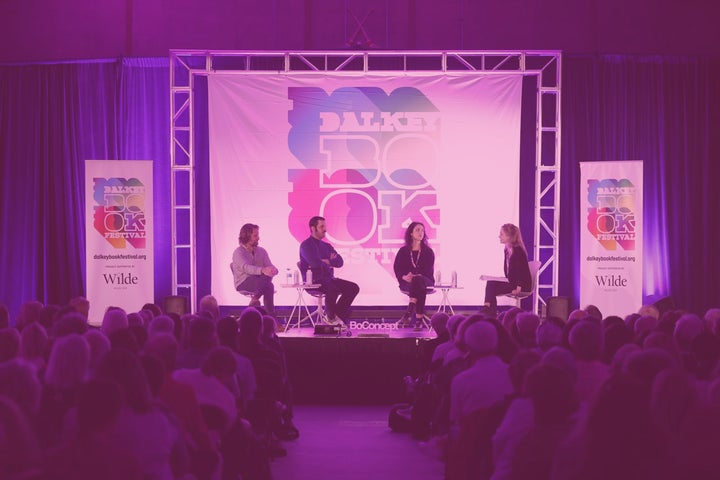 Fuzzy, pink lit view of a stage with 4 seated people in a discussion with a seated audience.