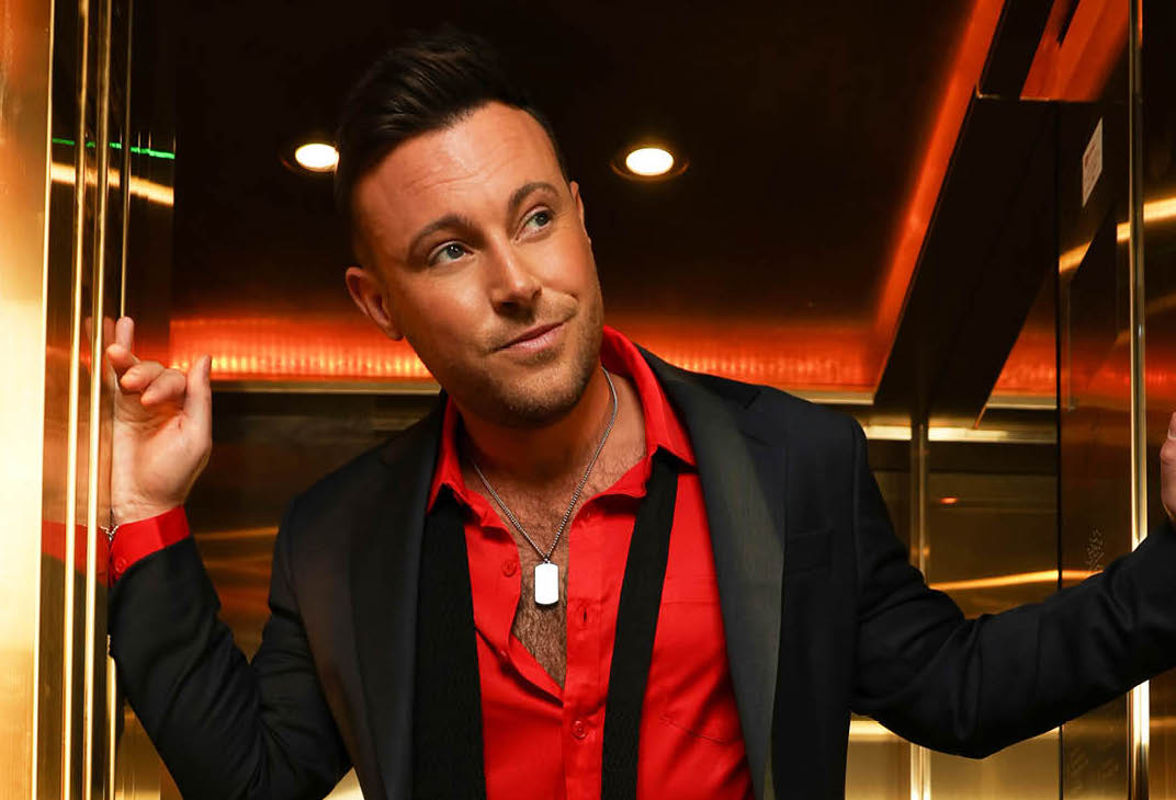 A man in a red open shirt and dark jacket appears to be looking out of the entrance of a small, gold lift, looking slightly bemused.