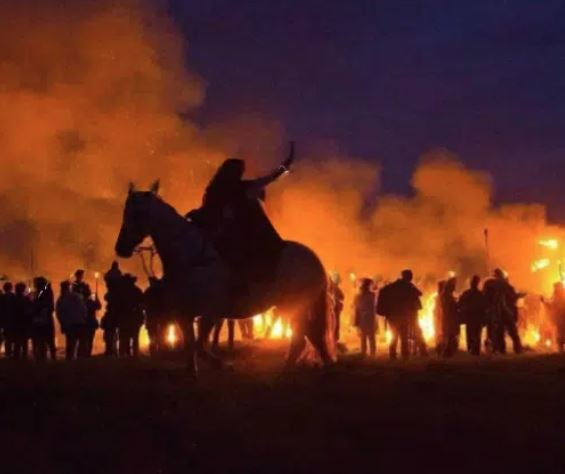 Person on a horse and line of figures at night silhouetted against flames and red smoke.
