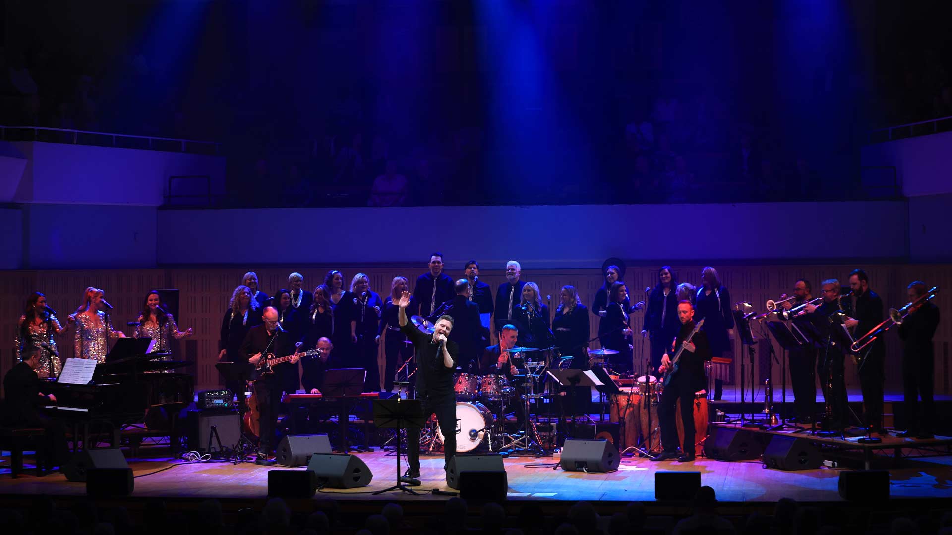 A man singing on a large stage with large band and choir behind him.