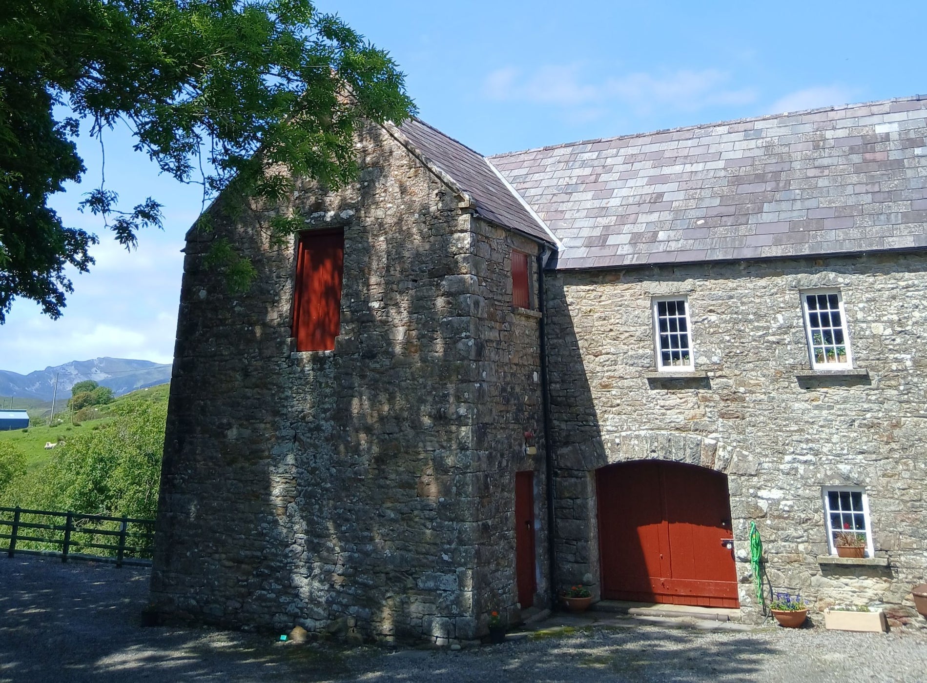 Exterior of a traditional stone corn mill with red doors with trees and hills in the background