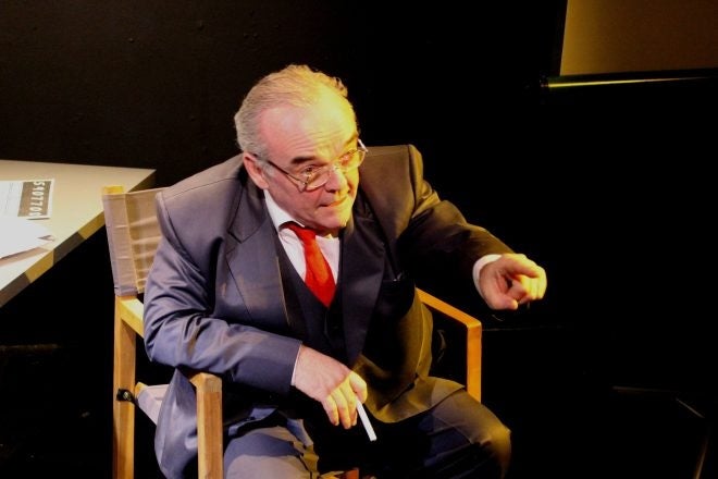 An older man in a suit is seated in a chair, talking and pointing with left finger