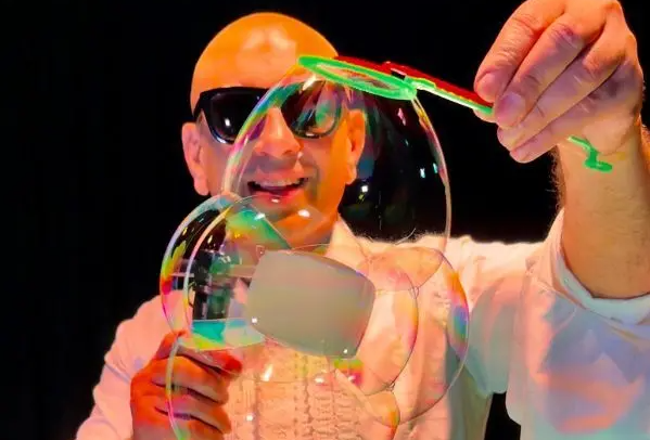 A smiling, bald man wearing sunglasses is holding an interlinked display of large bubbles, lit with orange light against black background.
