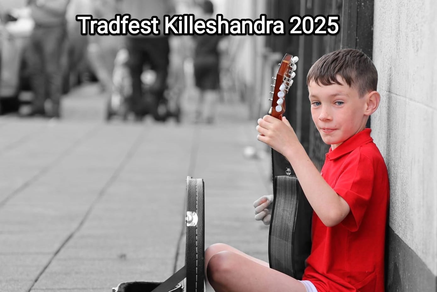 A young lad in bright red top is seated on a grey pavement leaning against a grey wall with a stringed instrument he is playing.