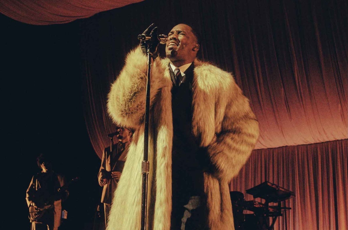 R&B superstar GIVĒON, a man in large, light coloured fur coat singing into a mic on a stand.
