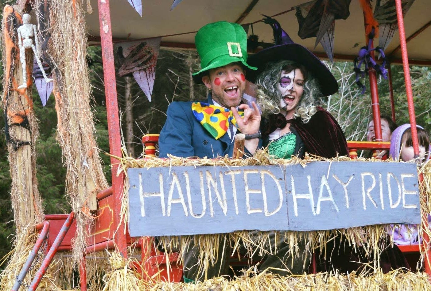 A person dressed as a witch beside a person dressed as a leprechaun on a trailer decorated with straw and halloween themed items.