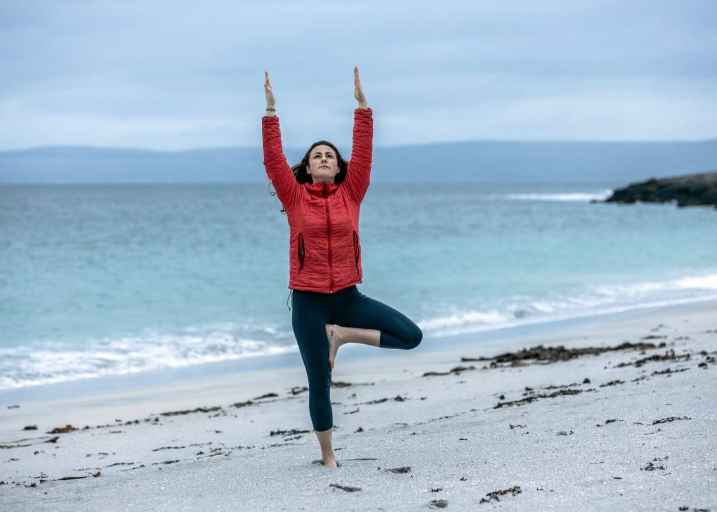 A woman doing a yoga tree pose with her hands in the air on the beach