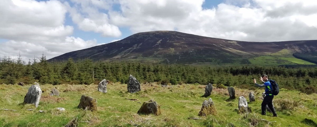 Man standing in front of standing stone circle with hands outstretched set against a wooded and mountain backdrop