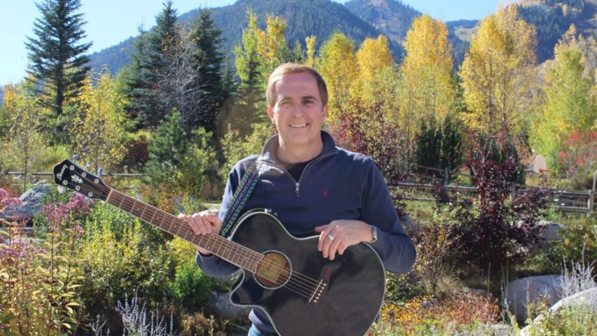 A smiling man with a guitar standing outdoors with trees in the background.