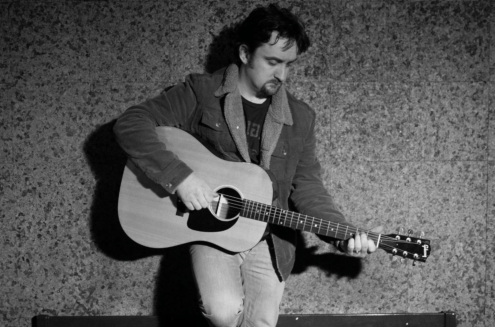 A man leaning against a wall playing a guitar looking down