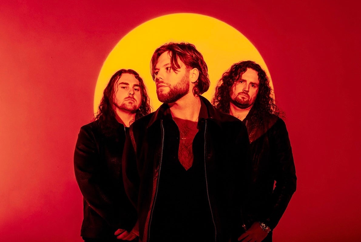 Rock band Florence Black, 3 moody looking men dressed in black with orange full sun background against red
