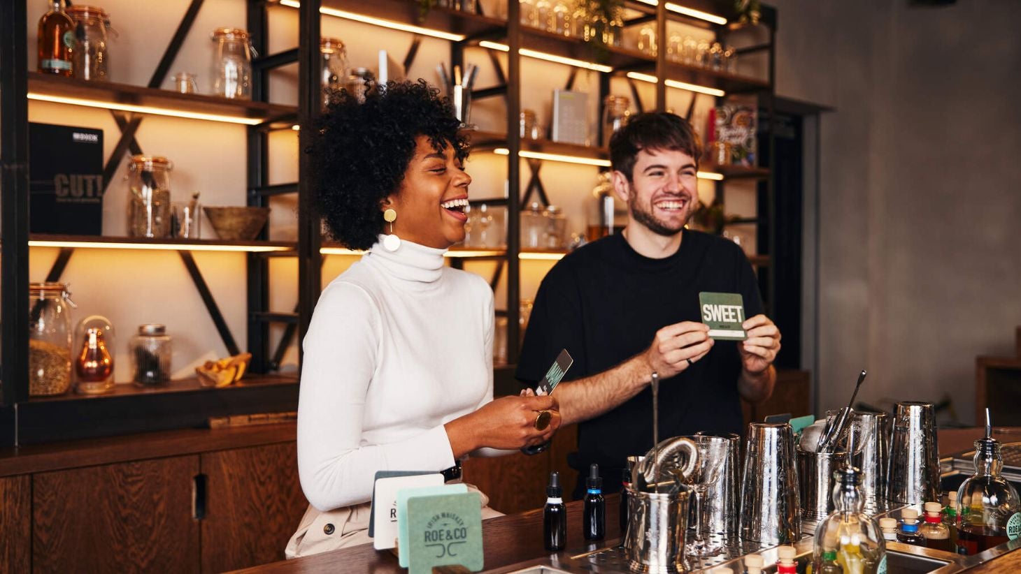 A woman and a man laughing behind the bar at Roe & Co Whiskey Tour as they explain something to tour goers.