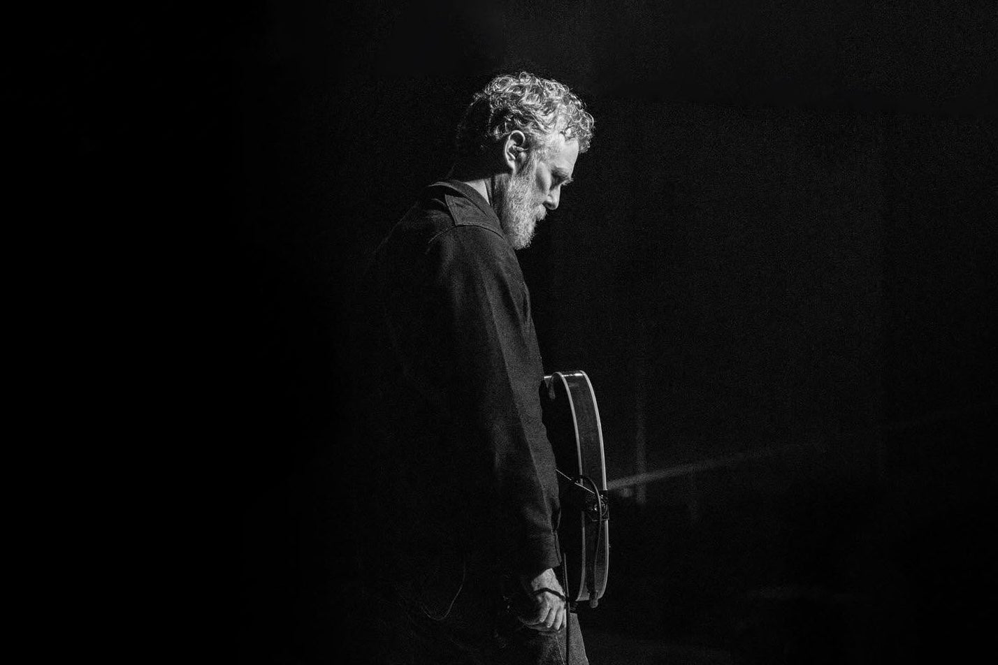 A dimly lit photo of man on a stage with a guitar, looking down.