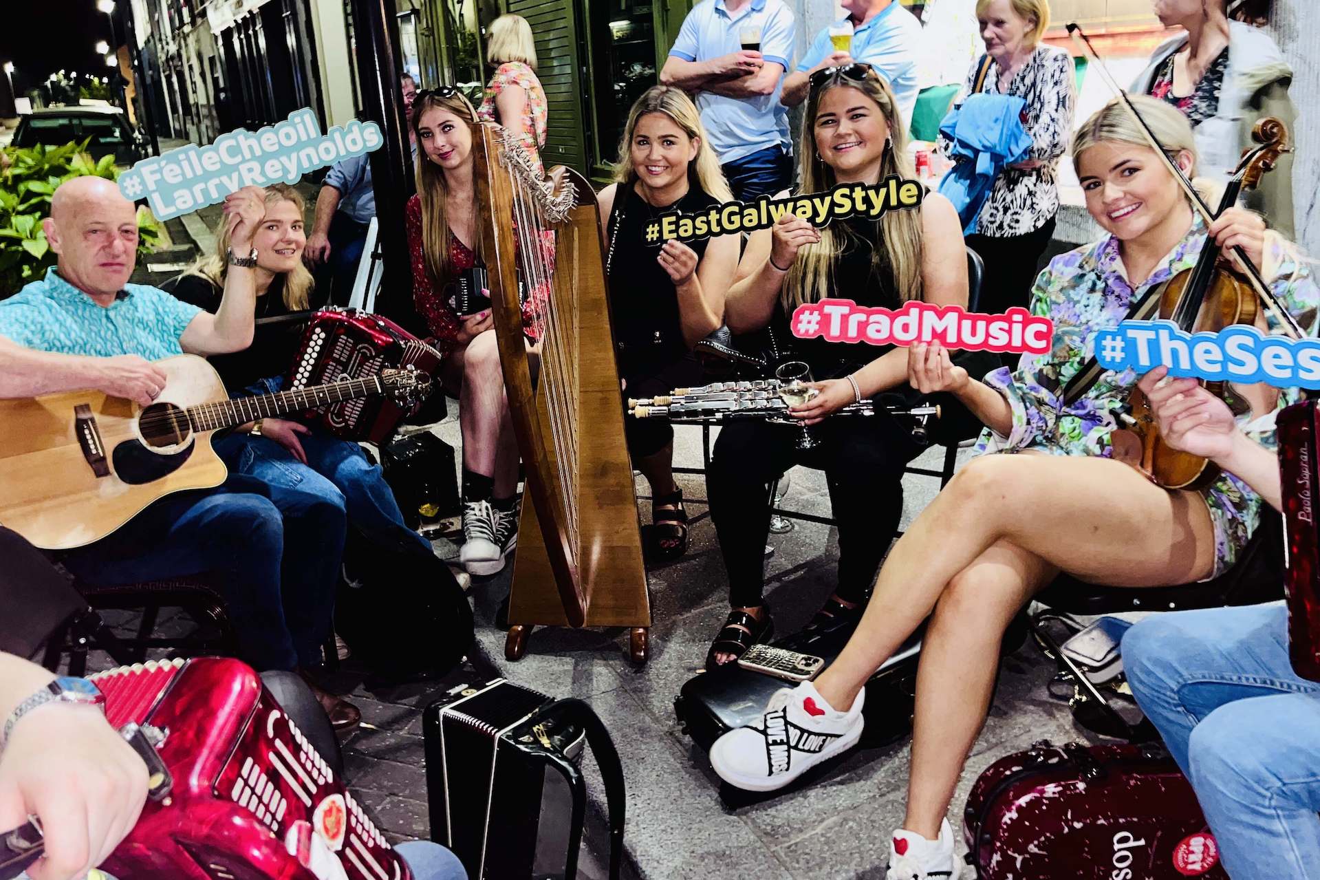 A group of musicians with instruments are seated in a close group on a pavement, some holding different coloured hash tag signs, smiling at the camera.