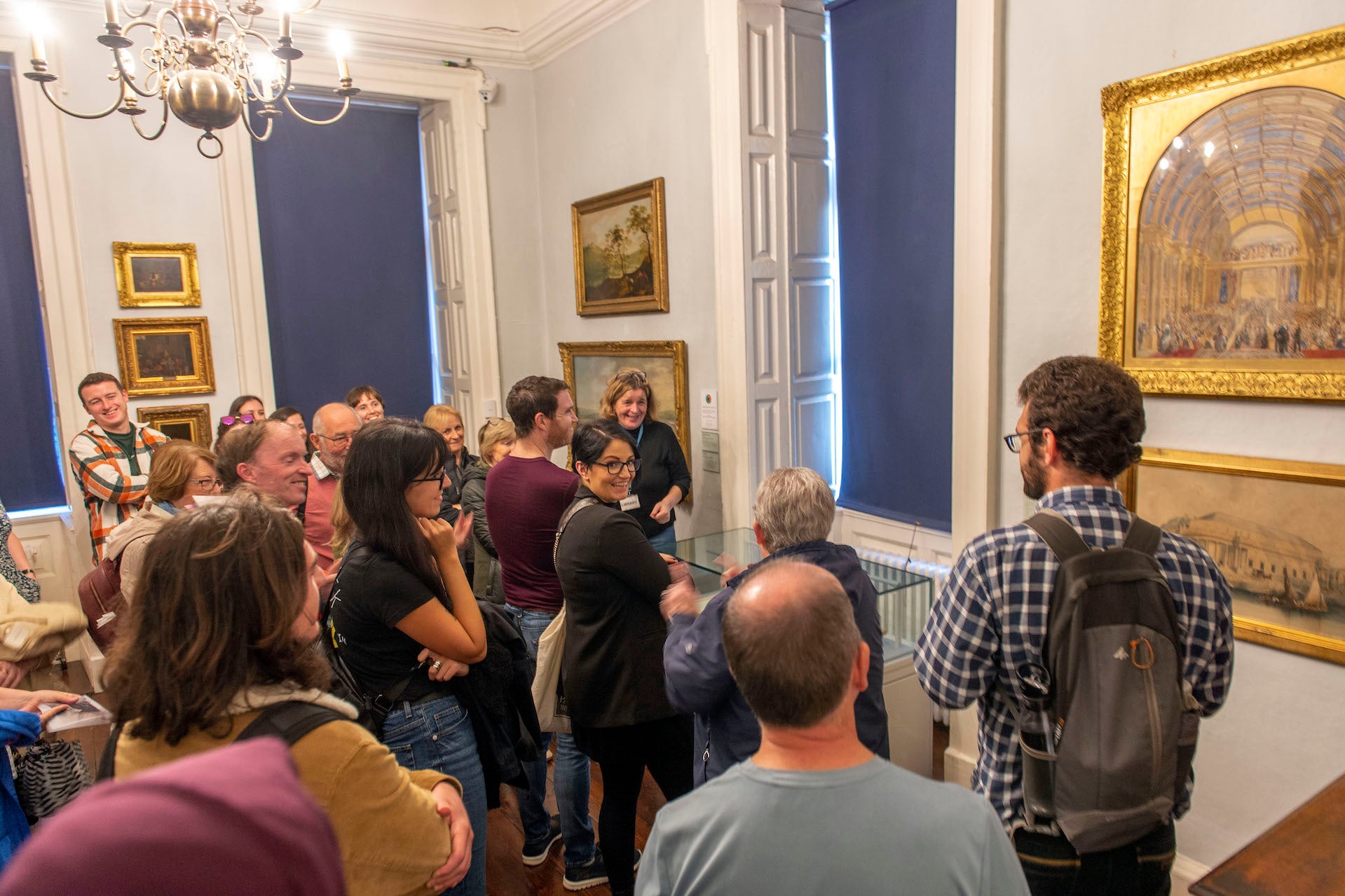 A group of people in a gallery are gathered round a small glass cabinet, listening to a tour guide speak.