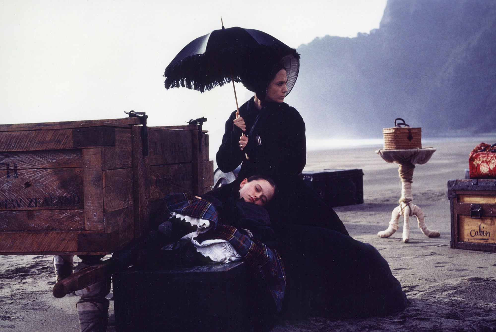 A girl resting against the lap of a woman with an umbrella. They are surrounded by luggage on a beach.