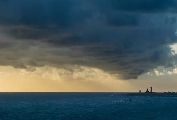 View the horizon over a flat, dark sea with dark clouds and a lighthouse in the distance.