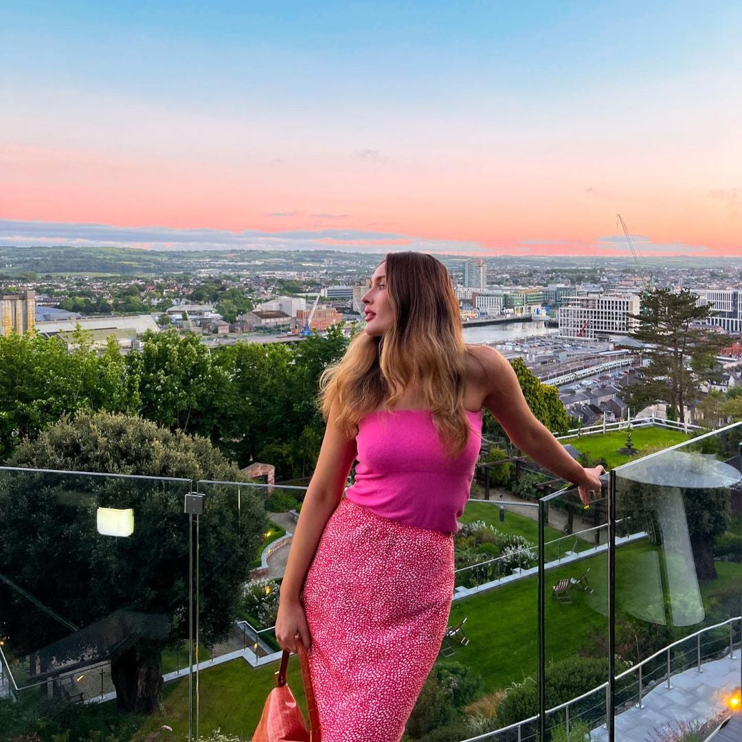 Woman dressed in pink top and skirt on hotel rooftop at sunset with Cork City behind her.