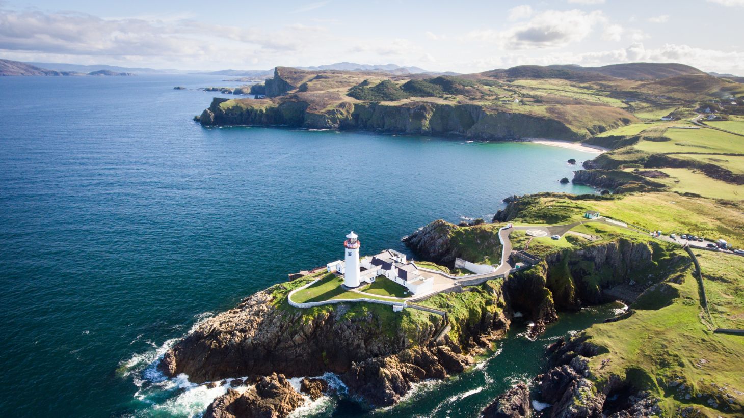 A lighthouse perched on a cliff with a backdrop of green fields and waves crashing against rocks