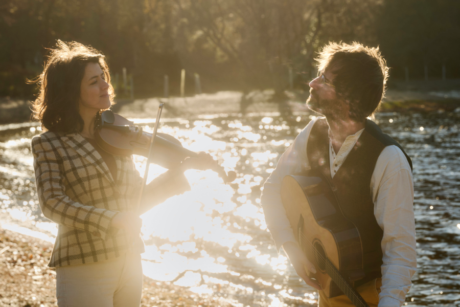 A woman playing a fiddle looking at a man playing a guitar, both standing outside with water and trees in the background.