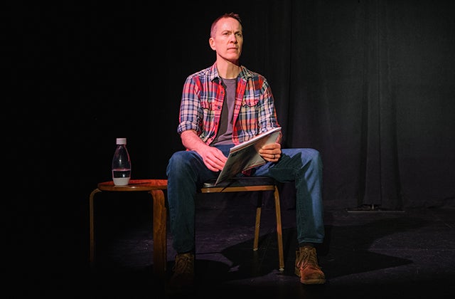 a man in checked shirt seated on a chair on stage