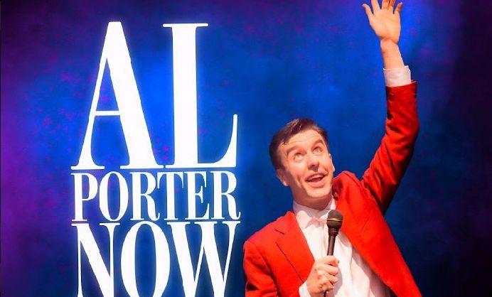 A man in red suit and white shirt and tie is holding a mic in one hand with the other up in the air over his head with a happy expression on his face, looking upwards, against blue background with white event text beside.