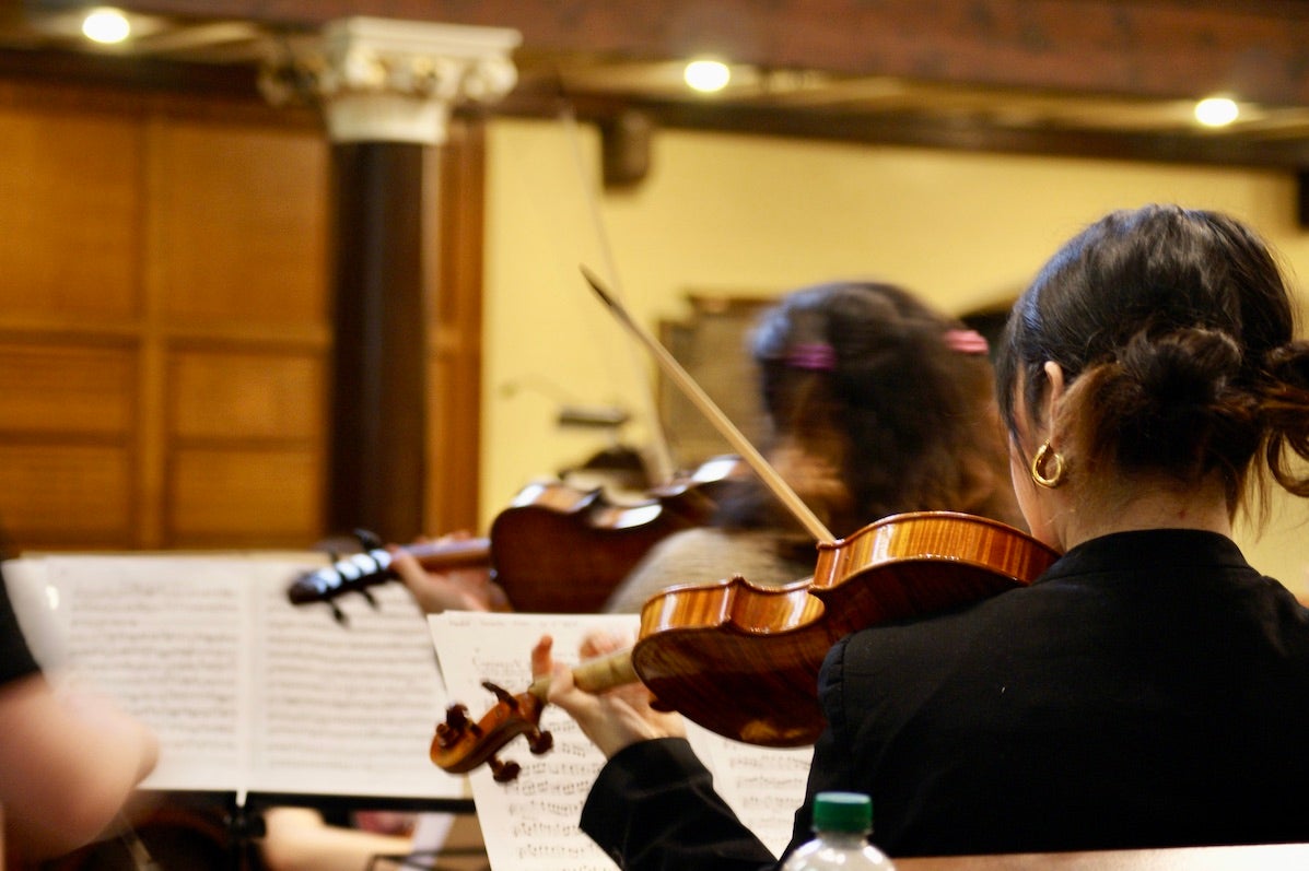 Review view of violinist playing with sheet music on stand in front and blurred other musicians.