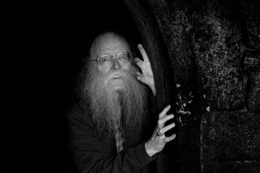 An older man in glasses with long busy beard, peering around a tree trunk in the dark.