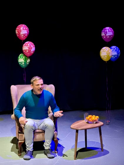 A man sits in an arm chair with Birthday balloons in the background