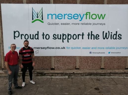 Two men standing in front of a Merseyflow sponsorship sign supporting the Wids