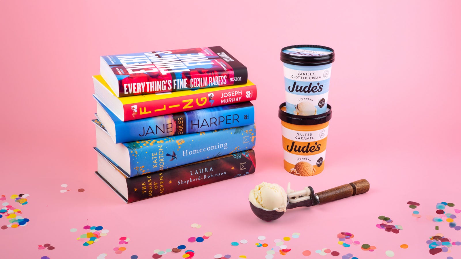 A stack of books next to two tubs of Judes Ice Cream stacked alongside an ice cream scoop They are in front of a pink background and surrounded by confetti