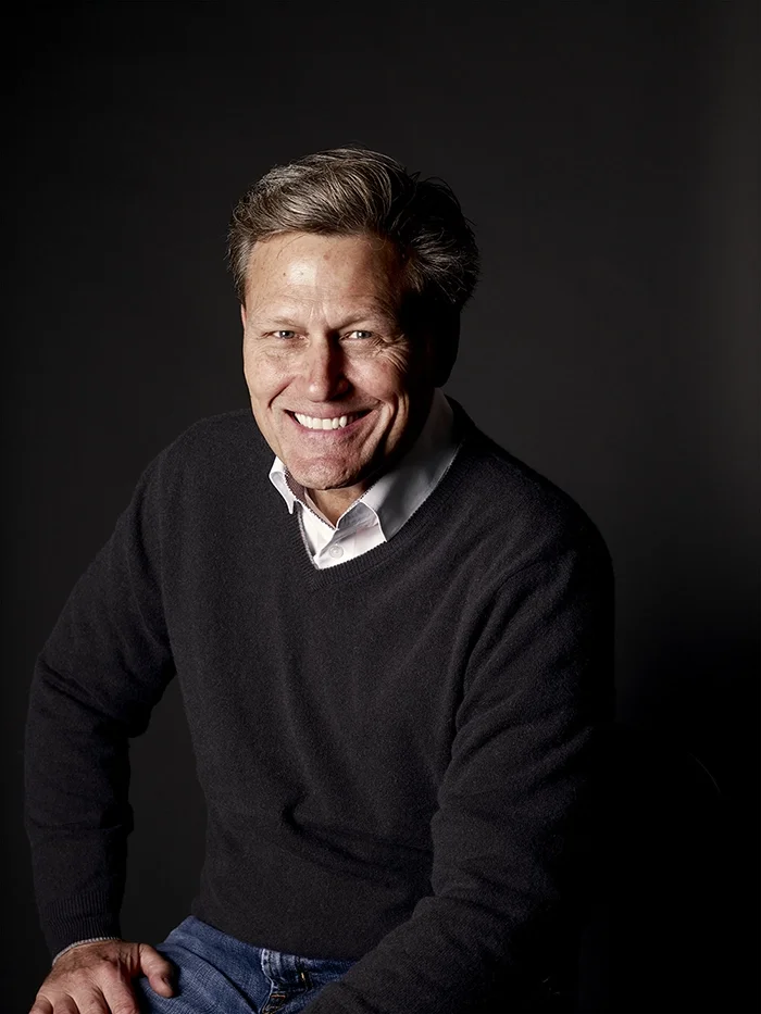 A photograph of David Baldacci wearing a grey jumper over a white shirt, and jeans. 